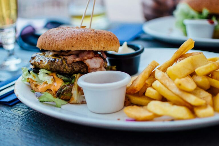 Der überraschende Rückzug der Restaurantkette Moto 59 vom Münsteraner Hafen wirft Fragen zur Standortwahl auf. Noch im Sommer 2024 war eine Neueröffnung am frisch entwickelten Hafenmarkt angekündigt. Doch stattdessen entschied sich Moto 59 gegen den Standort – offiziell wegen der starken Konkurrenz durch L’Osteria und The ASH. Ist dieses Argument stichhaltig? Und gilt der Hafen Münster generell als attraktiver für Gastronomiekonzepte als die Trauttmansdorffstraße (Standort der konkurrierenden Restaurants)? Eine Analyse der Gastronomie-Situation beleuchtet beide Standorte, die Konkurrenzlage sowie mögliche Verflechtungen der Betreiber. Gastronomie am Hafen Münster: Trendviertel mit hoher Frequenz und Preisen Der Hafen in Münster – oft „Kreativkai“ genannt – hat sich in den letzten zwei Jahrzehnten zu einer Top-Adresse für Gastronomie und Ausgehen entwickelt . Entlang des Hafenbeckens reihen sich über ein Dutzend Bars und Restaurants. Bereits 2017 zählte man rund 15 Gastronomiebetriebe mit Außengastronomie am Nordufer, die gemeinsam eine attraktive Ausgehmeile bilden. Insbesondere abends und am Wochenende pulsiert hier das Leben: Studenten, junge Berufstätige und Touristen genießen die vielfältigen Angebote am Wasser. Eine neu gestaltete Uferpromenade und die Mischung aus Kultur, Büros und Gastronomie sorgen für stetige Laufkundschaft und lebendige Atmosphäre. Allerdings hat die Beliebtheit ihren Preis. Das Hafenviertel ist durch Gentrifizierung geprägt – Mietpreise für Gewerbeflächen zählen zu den höchsten der Stadt. Für Büroflächen wurden bereits um 2010 Spitzenmieten von etwa 14 € pro Quadratmeter erzielt; für gastronomische Flächen in Premiumlage dürften ähnliche oder höhere Preise fällig sein. Zugleich ist die Konkurrenz am Hafen groß: Viele Lokale buhlen um ein ähnliches Publikum. Wer hier besteht, braucht ein überzeugendes Konzept, denn einfache Standard-Gastronomie geht in der Masse unter. Die Zielgruppe am Hafen – überwiegend ein trendbewusstes, junges Publikum – erwartet Vielfalt und Qualität. Die hohe Frequenz an Besuchern kann zwar volle Terrassen bedeuten, aber auch Lärm und Verkehrsaufkommen in den Abendstunden. Kurz: Der Hafen Münster bietet ein enormes Potenzial für Gastronomen, verlangt ihnen aber auch Innovationskraft und eine robuste Kalkulation ab. Trauttmansdorffstraße vs. Hafen: Zwei unterschiedliche Gastronomiestandorte Im Vergleich zum Hafenviertel präsentiert sich die Trauttmansdorffstraße als gänzlich anderer Standort. Hier, unweit des Preußen-Stadions und entlang des Dortmund-Ems-Kanals, hat sich jüngst ein neuer Gastronomieschwerpunkt entwickelt. Die Lage liegt am Rand der Innenstadt in einem Gewerbegebiet, das bislang weniger für Ausgehkultur bekannt war. Mit der Ansiedlung von L’Osteria und The ASH (Eröffnung im Oktober 2024) wird das Areal jedoch zum Anziehungspunkt. Beide Systemgastronomie-Riesen eröffneten nahezu zeitgleich an der Trauttmansdorffstraße 63–65 und investierten kräftig in ihre Standorte. The ASH etwa baute einen großzügigen Neubau („Freestander“) mit 180 Innenplätzen, großer Bar und 160 Terrassenplätzen, inklusive aufwändig gestalteter Piazza und Parkplätzen. Auch L’Osteria bietet hier viel Fläche und Parkmöglichkeiten – ein klares Signal, dass man auf Gäste mit Auto und Familien setzt, die bequem vor der Tür parken können. Passantenfrequenz und Umfeld unterscheiden sich deutlich vom Hafen: Während am Kreativkai viele Besucher spontan von Lokal zu Lokal ziehen, ist die Trauttmansdorffstraße eher ein Ziel-Standort. Gäste kommen gezielt, etwa für einen Familienabend, ein Steak-Dinner oder einen Pizza-Abend, oft mit dem Auto. Laufkundschaft vom Einkaufen oder Flanieren gibt es hier weniger, da es keine vergleichbare Kneipendichte ringsum gibt. Allerdings finden sich in der Nähe Freizeiteinrichtungen wie ein Bowling-Center und das Fußballstadion, was zu Stoßzeiten zusätzliches Publikum bringen kann. Mietpreise dürften in diesem peripheren Industriegebiet niedriger sein als in 1A-Lagen der Innenstadt – für große Ketten ein Vorteil, um auf großer Fläche zu expandieren. Die Eignung für Gastronomiekonzepte hängt hier stark vom Format ab: Große Erlebnisgastronomie mit Bekanntheitsgrad profitiert von der guten Erreichbarkeit und dem Platzangebot. Kleinere, unbekanntere Konzepte hingegen müssten ohne die Hafen-typische Laufkundschaft verstärkt in Marketing investieren, damit Gäste den Weg dorthin finden. Unterm Strich lässt sich kein Standort pauschal als „grundsätzlich attraktiver“ bewerten – es kommt auf das Konzept an. Der Hafen Münster garantiert Sichtbarkeit und hippe Umgebung, verlangt aber Alleinstellungsmerkmale, um sich gegen viele Mitbewerber abzuheben. Die Trauttmansdorffstraße bietet Expansionsfläche und weniger direkte Gastro-Nachbarn, dafür aber keinen eingebetteten Szenekiez. Attraktivität bedeutet hier also Unterschiedliches: Der Hafen lockt durch Flair und bestehende Publikumsmagneten, die Trauttmansdorffstraße durch Raum und Exklusivität im Umfeld. Konkurrenz durch L’Osteria und The ASH: Wie plausibel ist Moto 59s Argument? Die Betreiber von Moto 59 führen explizit die neue Konkurrenzsituation in Münster als Grund für ihren Rückzieher an. Tatsächlich hatten zwei Big Player – L’Osteria (italienische Pizza- und Pastakette) und The ASH (American Steakhouse/Bar-Konzept) – im selben Zeitraum massive Investitionen in Münster getätigt. Beide eröffneten Filialen, die konzeptionell zumindest teilweise mit Moto 59 überlappen: Moto 59 setzt auf ein Mix-Konzept aus Burgern, Pommes und Pasta für ein junges, trendiges Publikum. Genau diese Schnittmenge bedienen auch L’Osteria (italienische Pasta, Pizza in großer Portion für junge Leute) und The ASH (Burger und Bar-Ambiente für trendbewusste Gäste) in ihren neuen Niederlassungen. Die Big Magic Group aus Köln, Lizenzgeberin von Moto 59, sah darin offenbar ein ernstzunehmendes Risiko: Man fürchtete, dass die Millioneninvestitionen der Konkurrenz und deren starke Markenanziehung Moto 59 den Markteintritt in Münster erschweren würden. Schließlich verfügen L’Osteria und The ASH über eingespielte Konzepte, Marketingpower und eine bestehende Fanbasis – Faktoren, mit denen ein neuer Spieler erst einmal mithalten muss. Doch wie berechtigt ist dieses Argument? Einerseits erscheint es nachvollziehbar, dass Moto 59 nicht in einen Verdrängungswettbewerb mit zwei Gastronomie-Großprojekten gehen wollte. Die Konkurrenz belebt zwar das Geschäft, kann aber auch kleiner positionierten Konzepten schnell die Gäste abgraben, gerade wenn das Angebot ähnlich ist. Moto 59 hätte am Hafen zwar einen anderen Standort gehabt als L’Osteria und The ASH (die ja an der Trauttmansdorffstraße sitzen), doch in einer Stadt wie Münster bewegen sich Gäste innerhalb weniger Kilometer. Wer Lust auf Burger oder Pasta hat, hätte ab Ende 2024 die Wahl zwischen den etablierten Markenrestaurants mit großem Namen und dem neuen Moto 59. Aus Sicht der Moto 59-Macher war die Gefahr groß, dass viele die bekannten Konkurrenten vorziehen – sei es aus Neugier auf den neuen Steakhouse-Hotspot oder weil L’Osteria als Marke vertraut ist. Andererseits könnte man argumentieren, dass der Hafen als Standort Moto 59 auch Vorteile bot, die die Konkurrenten in ihrem Gewerbegebiet nicht haben: Laufkundschaft und Sichtbarkeit in einem populären Ausgehviertel. Einige Gastro-Fans in Münster reagierten verwundert, warum Moto 59 sich so sehr von der Konkurrenz einschüchtern ließ, statt es auf einen Versuch ankommen zu lassen. Immerhin wäre Moto 59 am Hafen derzeit der einzige Vertreter seines Konzepts, während an der Trauttmansdorffstraße gleich zwei große Restaurants direkt nebeneinander um Gäste konkurrieren. Dennoch darf man den Gesamtmarkt nicht vergessen: Münster kann nur eine gewisse Anzahl an neuen Restaurant-Plätzen verkraften. Die Marktsättigung im Segment „Burger/Pasta & Co.“ war durch die Neuankömmlinge vermutlich erreicht. Ohne einzigartige Positionierung liefen die Moto 59-Macher Gefahr, in einem schwierigen Umfeld zu starten. So gesehen ist das Rückzugs-Argument durchaus schlüssig – es spiegelt einen vorsichtigen strategischen Schritt, um Verluste zu vermeiden. Im Ergebnis steht das Hafen-Lokal bis auf Weiteres leer, doch der Projektentwickler sucht bereits nach einem neuen Mieter. Für den Standort an sich findet sich also vermutlich bald Ersatz, was andeutet: Nicht der Hafen ist das Problem, sondern die Kombination aus Konzept und Konkurrenz. Verflechtungen der Betreiber: Wie eng hängen Moto 59, L’Osteria und The ASH zusammen? Interessant ist bei genauerem Hinsehen, dass die beteiligten Marken und Personen in Münster teils persönlich und wirtschaftlich vernetzt sind. Ein möglicher leiser Faktor bei der Moto 59-Entscheidung könnte die Verbindung der Betreiber sein. So ist bekannt, dass Udo Hänold – der Betreiber mehrerer Moto 59-Filialen – früher für Kent Hahne gearbeitet hat. Kent Hahne wiederum ist kein Unbekannter in der Gastro-Branche: Er ist der Gründer und CEO von The ASH und steht somit hinter einem der neuen Großprojekte in Münster. Doch damit nicht genug der Überschneidungen. Hahne ist gleichzeitig der größte Franchisepartner von L’Osteria in Deutschland und betreibt aktuell 33 L’Osteria-Restaurants. Mit anderen Worten: Die beiden großen Konkurrenten, die Moto 59 abschreckten – L’Osteria und The ASH – stehen im Hintergrund zu einem guten Teil unter derselben unternehmerischen Führung. Welche Relevanz hat diese Verflechtung? Offiziell mag sie keine Rolle spielen, doch in der Gastronomiebranche sind Netzwerke und Beziehungen oft entscheidend. Man darf spekulieren, dass Udo Hänold die Expansionspläne seines früheren Chefs kannte und einschätzen konnte, welche Zugkraft dessen Betriebe in Münster entfalten würden. Eventuell wollte er keinen direkten Konkurrenzkampf mit Hahnes Konzepten eingehen – sei es aus Respekt vor dem ehemaligen Mentor oder aus nüchterner Einsicht in dessen Marktmacht. Dass Kent Hahne ausgerechnet beide neuen Restaurants in Münster (direkt oder indirekt) verantwortet, bedeutet auch: Marktstrategisch wurden hier keine Zufallsentscheidungen getroffen. Die parallele Eröffnung von The ASH und einer zweiten L’Osteria an der Trauttmansdorffstraße spricht für einen durchdachten Schachzug, die Münsteraner Gastrolandschaft gezielt zu besetzen. Moto 59 wäre als dritte ähnliche Marke im selben Zeitraum womöglich ins Hintertreffen geraten. Von wirtschaftlichen Verflechtungen im Sinne gemeinsamer Beteiligungen zwischen Moto 59 und den Konkurrenten ist zwar nichts bekannt – Moto 59 wird von der Kölner Big Magic Group als Franchisegeber vorangetrieben, während Hahnes Apeiron GmbH hinter The ASH steht und L’Osteria ein eigenes Franchise-System hat. Dennoch zeigt der Fall, wie personelle Überschneidungen indirekt Einfluss haben können. Die Entscheidung von Moto 59 mag offiziell mit Marktlogik begründet sein, doch die Hintergründe im Beziehungsgeflecht der Akteure liefern zusätzlichen Kontext: Ein starker Unternehmer dominierte bereits das Feld, ein aufstrebendes Konzept zog sich zurück. Fazit: Standortfrage und Konkurrenzdruck als Signal für Münsters Gastro-Markt Ist der Hafen Münster nun grundsätzlich attraktiver für Gastronomie als die Trauttmansdorffstraße? Für klassische Szenegastronomie und neue Bars sicherlich – hier bietet das Hafenviertel ein Publikum, das anderswo in der Stadt so nicht flaniert. Für große Systemgastronomie mit viel Platzbedarf kann dagegen ein weniger zentrumsnaher Standort mit Parkraum attraktiv sein, wie das Beispiel Trauttmansdorffstraße zeigt. Die Argumentation von Moto 59 wirkt vor diesem Hintergrund plausibel: Die Macher erkannten, dass weder der Standort Hafen noch die Ausweichadresse Trauttmansdorffstraße zum Erfolg geführt hätten, wenn gleichzeitig zwei große Konkurrenten einen Großteil der Zielgruppe abdecken. Statt am Hafen ins Rennen zu gehen und sich in unmittelbarer zeitlicher Nähe mit L’Osteria und The ASH messen zu müssen, zog Moto 59 die Reißleine. Für Münsters Gastronomie-Branche ist dieser Rückzug ein aufschlussreiches Signal. Er unterstreicht, dass Standortentscheidungen immer im Kontext des Gesamtmarktes gesehen werden müssen. Ein angesagtes Viertel allein garantiert keinen Erfolg, wenn das Konzept nicht einzigartig genug ist. Neue Restaurants – ob am Hafen oder anderswo – brauchen heute entweder eine klare Nische oder starke Alleinstellungsmerkmale, um sich gegen etablierte Mitbewerber zu behaupten. Die Episode zeigt auch, wie stark große Ketten die lokale Gastrolandschaft beeinflussen: Investieren sie Millionen und eröffnen gleich mehrfach, können kleinere Konzepte ins Wanken geraten, noch bevor der erste Burger über den Tresen gegangen ist. Münster bleibt ein attraktiver Markt, doch die Messlatte für neue Gastro-Konzepte liegt hoch – am Hafen genauso wie an der Trauttmansdorffstraße.