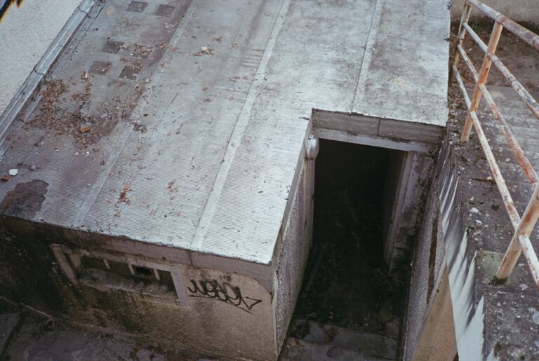 Bunker in Münster aus dem Zweiten Weltkrieg sind bis heute sichtbare Zeugnisse des Luftkriegs. Während des Zweiten Weltkriegs wurden in Münster zahlreiche Luftschutz-Bunker – sowohl oberirdische Hochbunker als auch unterirdische Anlagen – errichtet, um der Zivilbevölkerung Schutz vor Bombenangriffen zu bieten. Diese massiv aus Beton gebauten Schutzräume prägten einst das Stadtbild und einige von ihnen existieren noch heute. Der folgende Artikel gibt einen Überblick über die verschiedenen Bunker in Münster, ihre historische Nutzung im Zweiten Weltkrieg und ihren heutigen Zustand. Dabei werden oberirdische Bunker ebenso betrachtet wie unterirdische Anlagen und es wird beleuchtet, wie diese Relikte heute genutzt oder als Denkmäler erhalten werden. Historischer Hintergrund: Luftschutz in Münster im Zweiten Weltkrieg Münster war im Zweiten Weltkrieg als Gauhauptstadt von Westfalen-Nord und Sitz des Luftgaukommandos ein strategisch wichtiger Standort. Entsprechend wurde die Stadt häufig Ziel alliierter Luftangriffe. Zwischen 1940 und 1945 heulten in Münster über 1.100 Fliegeralarme, und die Bevölkerung suchte Schutz in den Bunkern. Insgesamt wurden in der Stadt rund zehn große Luftschutzbunker gebaut. Die meisten dieser Betonkolosse entstanden ab 1941/42, als der Luftkrieg an Intensität zunahm. Ihre Wände waren bis zu zwei Meter dick und die Decken etwa 1,4 Meter stark, um den Bombentreffern standzuhalten. Viele Münsteraner Bunker wurden von dem Architekten Edmund Scharf entworfen und verfügten über besondere Tarnmaßnahmen. Um aus der Luft weniger als militärische Ziele erkennbar zu sein, passte man das Erscheinungsbild der Bunker an zivile Gebäude oder historische Bauwerke an. So integrierten sich einige Bauten erstaunlich gut ins Stadtbild und sollten auf den ersten Blick nicht wie Bunker wirken. Dieser Aspekt der Tarnung zeigt sich an mehreren Beispielen in Münster. Oberirdische Bunker in Münster und ihre Tarnung Oberirdische Hochbunker (auch „Hochbauten“ genannt) prägten das damalige Stadtbild. Jeder dieser Bunker sah anders aus, da sie jeweils als zivile Gebäude getarnt geplant waren. Ein markantes Beispiel ist der Hochbunker an der Lazarettstraße: Dieser 1940/41 errichtete Bunker wurde äußerlich wie eine mittelalterliche Festung gestaltet, mit Zier-Rundtürmen, einer Zugangsbrücke und einem umliegenden Graben als Anspielung auf eine Burganlage. Wer den massivem Ziegelbau mit den bewachsenen Türmen sieht, könnte ihn für ein altes Fort halten – genau dieser Eindruck war beabsichtigt. Tatsächlich verbarg der Lazarettbunker hinter seinen Fassaden einen Schutzraum für bis zu 530 Menschen. Im Inneren ist die originale Ausstattung aus Kriegszeiten weitgehend erhalten, was ihn heute zu einem authentischen Zeitzeugnis macht. Ein weiteres Beispiel für kreative Tarnung war der Hochbunker an der Von-Kluck-Straße. Dieser fünfstöckige Bunker – der höchste in Münster – wurde 1942/43 auf dem Schulhof der Marienschule gebaut. Seine Fassade war als griechischer Tempel konzipiert, komplett mit Pfeiler-Andeutungen, um das militärische Aussehen zu verschleiern. Trotz dieser Tarnung erhielt der Von-Kluck-Bunker am 18. November 1944 einen Bombentreffer, überstand ihn jedoch ohne gravierende Schäden. Auf dem Dach dieses Hochbunkers befand sich sogar eine Flak-Stellung zur Luftverteidigung. Mit Platz für rund 2.000 Menschen zählte der Bunker zu den größten Schutzbauten der Stadt. Auch andere Hochbunker in Münster erhielten individuelle Gestaltungen: So sollte der Bunker am Hermannstadtweg ursprünglich ein Satteldach mit Wandmalereien im Heimatschutzstil bekommen. Zwar wurde die Tarn-Fassade dort nicht vollendet, doch auch dieser Betonbau hebt sich äußerlich von einem rein militärischen Aussehen ab. Insgesamt zeigt die Tarnung der Münsteraner Bunker, wie die NS-Bauplanung versuchte, die Luftschutzbauten in das städtische Umfeld einzupassen und gegnerische Bomber zu täuschen. Beispiele erhalten gebliebener Hochbunker und ihre heutige Nutzung Trotz Kriegsende und späterem Stadtumbau sind einige der großen Hochbunker Münsters bis heute erhalten. Drei ehemalige Bunker stehen sogar unter Denkmalschutz. Zu diesen gehört der bereits erwähnte Lazarettstraßen-Bunker, der sich im Besitz der Stadt Münster befindet und wegen seiner historischen Bedeutung als Baudenkmal gilt. Ein weiterer denkmalgeschützter Bunker ist der sogenannte Schützenhofbunker an der Wörthstraße im Südviertel. Dieser langgestreckte Bunker (ca. 61 m lang, 14,5 m breit, knapp 10 m hoch) wurde 1941/42 erbaut. Bei einem Bombentreffer im November 1944 wurde seine Decke beschädigt; die Druckwelle tötete 68 Menschen, die im Inneren Schutz gesucht hatten. Eine Gedenktafel an der Außenwand erinnert heute an diese Opfer. Nach dem Krieg erhielt der Schützenhofbunker eine neue Nutzung: In den vergangenen Jahren wurde der massive Bau privat umgebaut und enthält jetzt rund 38 Mietwohnungen auf etwa 1.600 Quadratmetern Fläche. Von außen ist die wuchtige Betonstruktur noch deutlich erkennbar, doch moderne Fenster und ein aufgesetztes Dachgeschoss zeigen die Wohnnutzung an. Dieser Umbau demonstriert eindrucksvoll, wie ein ehemaliger Luftschutzbunker in Münster erfolgreich in Wohnraum umgewandelt wurde – ohne seine geschichtliche Substanz ganz zu verlieren. Die Öffentlichkeit hat allerdings normalerweise keinen Zutritt, da es sich um Privatbesitz handelt. Ebenfalls noch vorhanden ist der Hochbunker am Hermannstadtweg (Geistviertel). Dieses 1941/42 errichtete Bauwerk mit ca. 2 Meter dicken Wänden umfasste einst 106 Räume. Anders als viele andere Bunker wurde er nicht abgerissen, sondern bereits seit Anfang der 1950er zivil genutzt. Heute befindet sich der Bunker Hermannstadtweg im Eigentum der Bundesanstalt für Immobilienaufgaben (BImA) und beherbergt diverse Künstlerateliers, Proberäume und Werkstätten. Unter anderem hat der Künstler Andreas Rosenthal dort seit rund 40 Jahren sein Atelier eingerichtet. Äußerlich ist der unverputzte Betonbau noch als solcher erkennbar, da das geplante Satteldach nie komplett fertiggestellt wurde. Trotzdem fügt sich der ehemalige Bunker inzwischen unauffällig ins Viertel ein, da er von Gewerbe und Kreativnutzung belebt wird. Ein ungewöhnliches Nachkriegs-Schicksal hatte ein Hochbunker im Stadtteil St. Mauritz. Dieser Bunker wurde nach 1945 in ein Schulgebäude umgewandelt und lange Zeit umgangssprachlich als „Bunkerschule“ bezeichnet. Ab 1949 diente er zunächst provisorisch als Volksschule, woraus später die Margaretenschule hervorging. Das massive Gebäude wurde teils entfestigt (entmilitarisiert) und mit Fenstern versehen, sodass es schließlich das Aussehen einer „etwas wehrhaften Unterrichtsstätte“ bekam. Bis heute wird es als Schulhaus genutzt und steht – ebenso wie der Lazarett- und der Schützenhofbunker – unter Denkmalschutz. Dieses Beispiel zeigt, wie flexibel die Stadt Münster nach dem Krieg mit den Bauten umging: Aus einem Ort des Schutzes vor Bomben wurde ein Ort des Lernens für Kinder. Nicht alle Bunker haben indes überdauert. Einige der ehemals zehn Anlagen wurden im Laufe der Jahrzehnte abgerissen oder stehen leer. Ein Hochbunker an der Emsstraße (Ecke Elbestraße, im Mauritzviertel), bekannt als „Emsbunker“, blieb viele Jahre ungenutzt und diente zeitweise als Lagerraum. Kürzlich wurde er von einem Investor gekauft, komplett zurückgebaut und abgerissen, um Platz für Neubauwohnungen zu schaffen. An seiner Stelle entstehen bis 2027 moderne Wohngebäude. Dieses Schicksal zeigt, dass die Zukunft mancher Bunker auch der städtebaulichen Entwicklung weichen muss, wenn keine schützende Denkmaleinstufung vorliegt. Unterirdische Bunker und Luftschutzkeller Neben den auffälligen Hochbunkern gab es in Münster auch unterirdische Luftschutzanlagen. Die bedeutendste darunter befindet sich unter dem Hauptbahnhof Münster. In den Jahren 1941/42 ließ die Deutsche Reichsbahn direkt unter dem Bahnhof einen großen Tiefbunker bauen, der rund 2.000 Menschen Schutz bieten sollte. Diese Anlage bestand aus ausgedehnten unterirdischen Räumen und starken Stahlbetonwänden. Trotz heftiger Bombardements auf die Bahnanlagen – allein 102 Luftangriffe der Alliierten galten dem Münsteraner Bahnhof und Güterbahnhof – überstand der Bahnhofsbunker den Krieg. Er wurde im Frühjahr 1945 von alliierten Truppen erobert, ohne dass es dabei zu Personenschäden kam. Bemerkenswerterweise ist dieser Bunker unter dem Hauptbahnhof auch heute noch erhalten. Während des Kalten Krieges wurde er sogar weiter ausgebaut und modernisiert, um im Falle eines Atomkriegs als Hilfskrankenhaus und Zivilschutzraum zu dienen. Hinter meterdicken Stahltüren verbergen sich dort Schleusen und Schutzräume, die jedoch der Öffentlichkeit normalerweise nicht zugänglich sind. Der Tiefbunker schlummert im Verborgenen unter dem Bahnhofsvorplatz – ein kaum sichtbares Relikt des Zweiten Weltkriegs mitten in der Stadt. Neben diesem großen Bahnhofsbunker gab es in Münster weitere unterirdische Schutzorte. Viele Wohnhäuser waren mit Luftschutzkellern ausgestattet, in die sich Bewohner bei Alarm zurückzogen. Bis heute finden sich an einigen älteren Häuserfassaden noch eingemeißelte oder gemalte Luftschutzkeller-Pfeile, die den Weg zu diesen Kellern wiesen. Solche Pfeile – kleine Dreiecke oder Pfeilsymbole mit der Aufschrift "LS-Raum" oder ähnliches – sind verbliebene Spuren der Luftschutzmaßnahmen im Stadtbild. Auch die Tunnelanlagen der städtischen Befestigungen (etwa am Zwinger oder in ehemaligen Kasernen) wurden zeitweise als Bombenschutzräume genutzt, soweit es möglich war. Ein Beispiel für kleinere spezielle Bauten der Reichsbahn sind sogenannte Rundbunker: Am Rande des Güterbahnhofs in Münster standen mehrere runde Betonbunker, die dem Schutz von Bahnpersonal dienten. Einige Überreste solcher Rundbunker sind ebenfalls erhalten geblieben, gut getarnt im Bewuchs oder integriert in heutige Nutzungen. Vom Kriegsrelikt zum Denkmal: Heutiger Umgang und Gedenken Ein großer Teil der Bunker in Münster ist heute entweder einer neuen Nutzung zugeführt oder als Mahnmal erhalten. Drei ehemalige Luftschutzbunker – der Lazarettbunker, der Schützenhofbunker und die Bunkerschule St. Mauritz – stehen auf der städtischen Denkmalliste und werden als historische Bauwerke anerkannt. Dies bedeutet, dass ihr Erhalt im öffentlichen Interesse liegt. Die Stadt Münster bemüht sich, diese Relikte des Zweiten Weltkriegs in das kollektive Gedächtnis zu rücken. So wurden etwa im Rahmen des bundesweiten Tags des offenen Denkmals am 10. September 2023 erstmals seit langer Zeit Bunker der Öffentlichkeit zugänglich gemacht. An diesem Tag konnten interessierte Bürger den Hochbunker an der Lazarettstraße und den Schützenhofbunker von innen besichtigen – ein Angebot, das regen Zuspruch fand. Solche Veranstaltungen sollen bewusst machen, welche bedrückende Zeitgeschichte in den massiven Mauern steckt. Auch durch Gedenktafeln wird an die Geschichte der Bunker erinnert. Der Schützenhofbunker trägt, wie erwähnt, eine Tafel für die 68 Opfer des Bombentreffers von 1944. Am Lazarettbunker wiederum informieren Hinweisschilder über die außergewöhnliche Architektur und die Tarnung als Burg. Initiativen von Heimatvereinen und Geschichtsinteressierten bieten außerdem gelegentlich Führungen zu den Bunkerstandorten an, um die Erinnerung wachzuhalten. In Publikationen und Online-Archiven werden Fotos und Zeitzeugenberichte zu den Münsteraner Bunkern gesammelt, damit dieses Kapitel der Stadtgeschichte nicht in Vergessenheit gerät. Schließlich stellt sich die Frage nach der Zukunft dieser Bauten. Einige, wie der Emsbunker, weichen der Stadtentwicklung und verschwinden zugunsten neuer Gebäude. Andere wie der Bunker Hermannstadtweg haben sich längst in den Alltag integriert und erfüllen eine neue Funktion. Wieder andere stehen zwar leer, sind aber als Mahnmale erhalten. So sind die Bunker in Münster heute vielgestaltig: Sie dienen als Wohnungen, Lager, Ateliers oder Schulgebäude, oder sie ruhen still und erinnern an die Schrecken des Krieges. Fazit: Die Bunker in Münster aus dem Zweiten Weltkrieg erzählen von Angst, Schutzsuche und Überlebenskampf während der Luftangriffe. Oberirdisch wie unterirdisch waren sie für Tausende Münsteraner lebensrettend. Heute sind sie stumme Zeugen der Geschichte – teils vergessen im Stadtbild, teils bewusst konserviert. Indem man diese Betonrelikte bewahrt und ihrem Schicksal gedenkt, hält Münster die Erinnerung an die dunklen Zeiten des Bombenkriegs lebendig und mahnt zugleich zum Frieden. Quellen: Stadtarchiv Münster, Zeitungsberichte und Denkmaldatenbanken über Münsteraner Bunker, sowie lokale Initiativen und historische Dokumentationen.