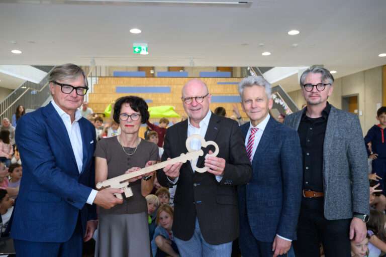 Am Montag, 22. September 2025, hat die Stadt Münster den Erweiterungsbau der Grundschule Wolbeck-Nord feierlich eingeweiht. Mit einer symbolischen Schlüsselübergabe an Schulleiterin Christiane Kröger wurde das moderne Gebäude offiziell übergeben. Oberbürgermeister Markus Lewe, Stadtdirektor Thomas Paal und Vertreter des Bauplanungsbüros Ripperda sowie der Baufirma Mod21 waren bei der Eröffnung vor Ort. Erweiterungsbau Grundschule Wolbeck-Nord als Antwort auf steigende Schülerzahlen Der neue Bau ist Teil der Schulbauoffensive der Stadt Münster. Besonders im Südosten und in Hiltrup wächst die Zahl der Schülerinnen und Schüler, während andere Stadtbezirke stagnieren. Mit dem Erweiterungsbau reagiert die Stadt auf diesen steigenden Bedarf und schafft zusätzliche Kapazitäten für Unterricht und Betreuung. Seit Beginn des neuen Schuljahres wird der Neubau bereits genutzt, wodurch die Grundschule nun dreizügig geführt werden kann. Rekordbauzeit dank modularer Bauweise Ein besonderes Merkmal des Projekts ist die extrem kurze Bauzeit. Nur fünf Monate vergingen vom Start bis zur Fertigstellung der Klassenzimmer. Möglich wurde dies durch die modulare Bauweise mit vorgefertigten Holzelementen. Rund 5,4 Millionen Euro investierte die Stadt Münster in den zweigeschossigen Neubau am Grenkuhlenweg. „Mit dem Erweiterungsbau der Grundschule Wolbeck-Nord schaffen wir ein Gebäude, das pädagogische Qualität, Nachhaltigkeit und kurze Bauzeit überzeugend verbindet“, erklärte Oberbürgermeister Markus Lewe bei der Eröffnung. Moderne Lernräume und flexible Nutzung Das neue Gebäude bietet vier Klassenräume, zwei Differenzierungsräume, drei Betreuungsräume sowie einen Mehrzweckraum. Für die Verwaltung wurden zwei Büros, eine Teamstation und eine Teeküche eingerichtet. Breite Flure mit Aufenthaltsnischen ermöglichen den Kindern zudem, auch außerhalb der Klassenräume zu lernen oder sich auszutauschen. Schulleiterin Christiane Kröger betonte, dass der Neubau weit mehr sei als ein zusätzlicher Platzgewinn: „Unsere Kinder nutzen die Räume, um individuell zu lernen, sich zu bewegen, kreativ zu gestalten und Neues zu entdecken. Das Gebäude wird bereits jetzt als vertrauter Lern- und Lebensraum angenommen.“ Nachhaltigkeit als zentrales Element Auch ökologisch setzt der Erweiterungsbau der Grundschule Wolbeck-Nord Maßstäbe. Die Module bestehen aus zertifiziertem Holz. Auf dem Dach sorgt eine Photovoltaikanlage mit 40 Kilowatt-Peak Leistung künftig für die Einsparung von rund zehn Tonnen CO₂ pro Jahr. Zusätzlich tragen eine Dachbegrünung und eine bepflanzte Ostfassade zur Verbesserung des Mikroklimas bei. Planung und Umsetzung Die Projektsteuerung lag beim Amt für Immobilienmanagement der Stadt Münster. Die Planung übernahm das Bauplanungs- und Ingenieurbüro Ripperda aus Lingen. Für die Umsetzung war das Unternehmen Mod21, Tochter der Erbud-Gruppe, verantwortlich. Durch die enge Zusammenarbeit aller Beteiligten konnte das Projekt in kurzer Zeit und im vorgesehenen Kostenrahmen realisiert werden.